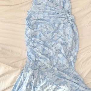 Ljusblå långklänning med blommönster - Superfin ljusblå långklänning som kan bäras offshoulder  med  blommigt mönster. Klänningen har snygg rynkning längs sidorna och en figurnära passform som framhäver siluetten. Testat en gång. Köpte precis för 950kr.