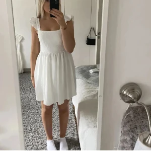 Vit kortklänning med volangärmar - Superfin vit kortklänning med volangärmar och fyrkantig ringning. Smockad rygg för skön passform och lätt utsvängd kjol. Perfekt för sommarens alla tillfällen. Klänningen är i ett tunt, luftigt material och har en söt, feminin look. ( Andvänder bilder från tjejen innan mej)