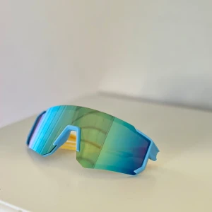 Sportiga barn solglasögon - Säljer ett par futuristiska sportglasögon med spegelblå/gröna linser, blå båge och gula skalmar. De har en skarp, kantig design och ger riktigt cool vibe. Perfekta för dig som vill sticka ut på cykeln eller löprundan😎