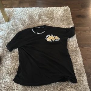 Svart Palm Angels t-shirt med tryck - Svart t-shirt från Palm Angels med korta ärmar och rund hals. Snyggt tryck med björnmotiv och Palm Angels-logga på bröstet samt textdetalj runt halsen. Skön passform och streetwear-vibe, perfekt för dig som gillar trendiga märken. Den är large fast tight