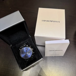 Emporio Armani svart/blå klocka - Stilren klocka från Emporio Armani med svart mesh-armband i metall och blå urtavla. Klockan har kronograf, datumvisning och silverfärgade detaljer. Levereras i originalbox med manual. Perfekt accessoar för dig som gillar klassisk design med modern touch.