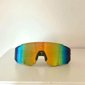 Snygga sportiga solglasögon  - Säljer ett par futuristiska sportglasögon med spegelglas i regnbågsfärger som skiftar mellan blått, grönt och gult. De har en bred, kantig form och svarta skalmar med coola utskärningar😎Perfekta för dig som vill sticka ut på träningen eller cykelturen💪