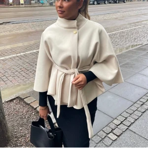 Beige poncho från Missay - Superfin beige poncho från Missay med knytbälte i midjan och guldknapp vid halsen. Den är bara använd en gång, men har fått lite smink på kragen (se bild 3) men inget man märker, och går nog bort i tvätten.