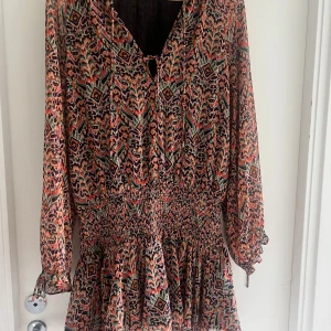 Mönstrad kortklänning från Sfera - Färgstark kortklänning från Sfera med unikt mönster i svart, orange, grön och rosa. Klänningen har långa ärmar med resår vid handlederna, smockad midja och v-ringning med knyt. Perfekt för dig som gillar statement-plagg och coola detaljer.