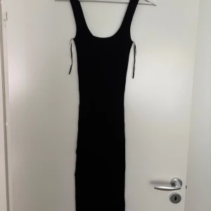 Svart ribbad långklänning Gina Tricot - Svart långklänning från Gina Tricot i ribbat material. Klänningen är ärmlös med breda axelband och har en tight passform som framhäver figuren. Enkel och stilren design med rundad ringning och knytband i sidorna.