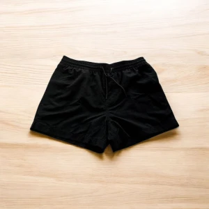 Svarta träningsshorts med snörning - Svarta shorts fran Visual Clothing by MQ med elastisk midja froch snörning framtill. De har en bakficka med knapp och meshfoder på insidan, vilket gör dem perfekta för träning eller bad. Klassisk och enkel design som passar till det mesta.