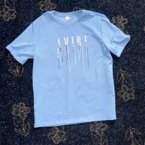Ljusblå AMIRI t-shirt - Snygg ljusblå t-shirt från AMIRI med coolt vitt tryck på bröstet där bokstäverna ser ut att smälta neråt. Klassisk passform och rund halsringning, perfekt för en stilren och trendig look.