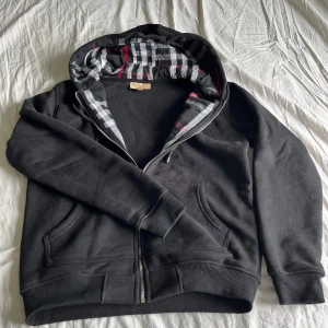 Grå zip-hoodie från burberry - Svart hoodie från Burberry med klassiskt rutigt foder i huvan i svart, vitt och rött. Tröjan har dragkedja framtill, två fickor och ribbade muddar. Perfekt för dig som gillar streetstyle och vill ha något bekvämt men ändå snyggt. Pris kan diskuteras vid snabb affär.