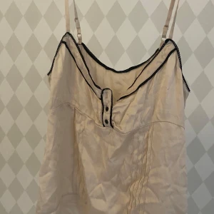 Beige linnetopp med svarta detaljer - Säljer en beige linnetopp med tunna axelband och svarta kantdetaljer. Toppen har knappar framtill och dekorativa sömmar längs nederkanten och framtill. Perfekt för en chill sommarlook eller att styla lager på lager. Kan gå ner i pris vid en snabb affär💛💛