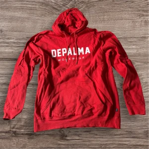 Röd hoodie från Depalma Workwear XL - Riktigt snygg röd hoodie från Depalma Workwear i storlek XL. Tröjan har vit logga framtill, klassisk känguruficka och huva med snörning. Mjuk insida och långa ärmar, perfekt för kyliga dagar. Passar dig som gillar streetwear och enkel stil.