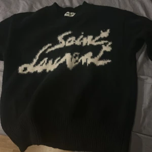 Svart stickad Saint Laurent tröja - ALDRIG ANVÄND! Svart stickad tröja från Saint Laurent med vit, suddig logotyp framtill. Tröjan har rund halsringning och ribbade muddar vid ärmslut och nederkant. Perfekt för dig som vill ha en statement piece med lyxig känsla.