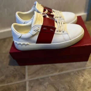 Valentino Garavani Open sneakers vit/röd - Snygga Valentino Garavani Open sneakers i vitt skinn med bred röd läderdetalj över vristen och ikoniska nitar baktill. Klassisk rund tå, platt sula och vita snören. Perfekta för dig som vill sticka ut med exklusiva detaljer och stilren design.