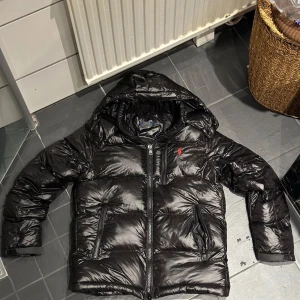 Polo Ralph Lauren jacka - Snygg svart dunjacka från Polo Ralph Lauren med huva och dragkedja. Jackan har en röd broderad logga på bröstet och är perfekt för kyliga dagar. Den är långärmad och har en glansig finish.