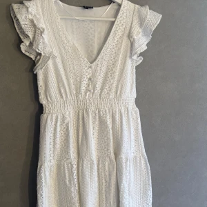 Vit spetsklänning med volangärmar - Superfin vit kortklänning i spets med v-ringning, volangärmar och smock i midjan. Klänningen har dekorativa knappar framtill och ett romantiskt mönster i spetsen. Perfekt för sommarens alla tillfällen!