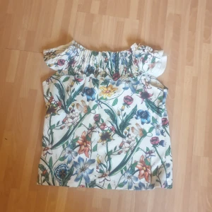 Blommig offshoulder topp från Zara - Superfin offshoulder topp från Zara Trafaluc med färgglatt blommönster på vit botten. Toppen har volang vid axlarna och är ärmlös, perfekt för varma dagar. Materialet är mjukt och lätt, vilket gör den skön att bära hela sommaren.