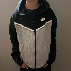 Grå och svart huvtröja från Nike - Säljer en snygg grå och svart huvtröja från Nike med dragkedja och huva. Tröjan har en vit randdetalj över bröstet och en praktisk ficka med dragkedja på ärmen. Perfekt för en sportig look!