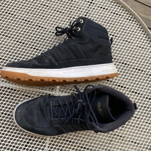 Svarta höga sneakers från Adidas - Snygga svarta höga sneakers från Adidas med klassiska tre ränder på sidan, gulddetaljer vid snörningen och grov gummisula i brunt. Ovandel i mocka och textil, perfekt för dig som gillar streetstyle och vill ha något som sticker ut.