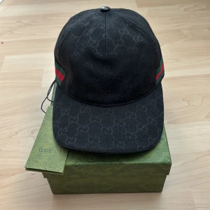 Svart Gucci keps canvas - Snygg svart keps från Gucci med klassiskt GG-monogram över hela kepsen och gröna/röda ränder på sidorna. Justerbar rem baktill i skinn med präglad Gucci-logga. Perfekt accessoar för att lyfta din outfit med lyxig känsla. Box ingår ej!!!!
