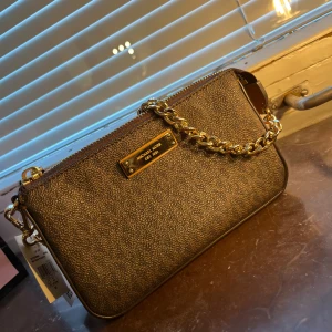 Brun axelväska från Michael Kors - Snygg brun axelväska från Michael Kors med guldfärgad kedja och dragkedja upptill. Väskan har en stilren rektangulär form och är tillverkad i skinnliknande material. Invändigt finns flera fack och Michael Kors-logga på fodret. Perfekt för att lyfta din outfit. I super bra skick förutom nån fläck i botten av nagelim, Syns inte mycket 500 eller ge bud