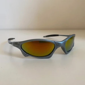 Oakley  - Oakley X-Metal solglasögon med futuristisk, sportig design. Bågarna är i matt silverfärgad metall och glasen har spegelreflex i gult och orange. Smal passform med svarta detaljer på skalmarna. Perfekta för dig som vill sticka ut med en edgy stil. Perfekta till sommaren. 