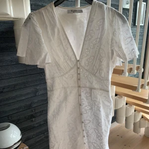 Vit broderad kortklänning från NA-KD - Superfin vit kortklänning från NA-KD med broderade detaljer och v-ringning. Klänningen har korta ärmar, knappar framtill och ett romantiskt mönster. Perfekt för sommarens alla tillfällen och ger en fräsch och luftig känsla.