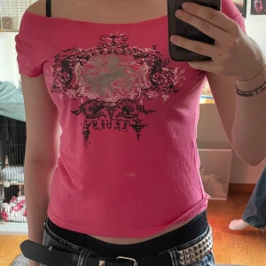 Rosa offshoulder topp med tryck - Snygg rosa offshoulder topp med svart och silverfärgat tryck framtill i barockstil. Modellen är croppad och sitter tight, perfekt för dig som gillar en edgy vibe. Tunn och mjuk bomullskänsla, passar till både jeans och kjol.