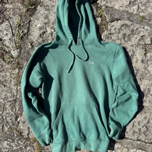 Grön hoodie från Vans med huva - Snygg grön hoodie från Vans med broderad logga på bröstet. Klassisk modell med huva, snörning och känguruficka framtill. Streetstyle. Av bomull. Mått: Chest( A): 54,5cm , Length(B):68,5cm, Arms: 62cm