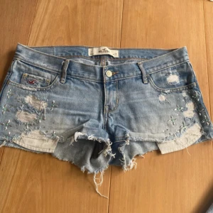 Lågmidjade ljusblå jeansshorts - Ljusblå jeansshorts från Hollister med råa kanter, slitningar och broderade detaljer med små pärlor framtill. Klassisk femficksmodell med låg midja och snyggt sliten look. Perfekta för sommaren och ger en avslappnad vibe.