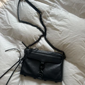 Svart axelväska från Rebecca Minkoff - Hej säljer en Snygg svart axelväska i skinn från Rebecca Minkoff med kedjerem och coola metalldetaljer framtill. Väskan har en stilren design med lock och dragkedja. 
