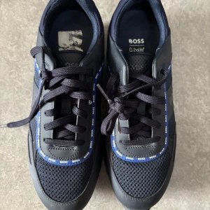 Marinblå sneakers från BOSS - Snygga marinblå sneakers från BOSS med svarta och blå detaljer samt märkets logga längs snörningen. Ovandelen är i mesh och syntetmaterial för en sportig look. Skorna har rund tå och platt sula.