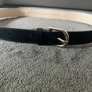 Svart bälte från H&M med silverfärgat spänne - Stilrent svart bälte från H&M i storlek S med ett klassiskt silverfärgat spänne. Bältet är gjort i skinnimitation med slät yta och diskreta sömmar längs kanterna. Perfekt accessoar för att lyfta din outfit med en clean och tidlös look.