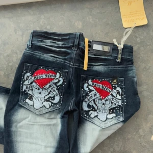 Miss Uwa jeans med döskalle och hjärta - ‼️kan köpas den 8 aug‼️Mörkblå jeans med slitna detaljer och blekta partier. På bakfickorna finns stora broderier med döskalle, hjärta och text, dekorerade med pärlor. Framtill finns liknande broderi på ena benet. Klassisk femficksmodell med låg midja och bootcut-snitt. Kan skicka mått när jag är hemma💗men storlek w26! Prislappen är borttagen men har den kvar