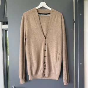 Beige kofta från Tiger of Sweden - Snygg beige kofta från Tiger of Sweden i mjuk ullmix. Koftan har v-ringning, knappar framtill och två fickor. Långärmad med ribbade muddar och axelparti. Perfekt för lager-på-lager och stilren look.