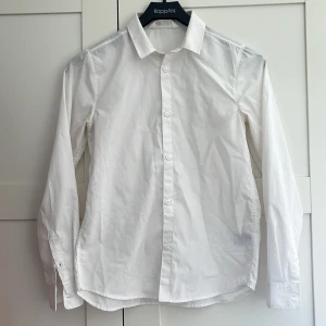 Vit skjorta från H&M, stl 152 - Klassisk vit skjorta från H&M i storlek 152. Skjortan har lång ärm, knappar framtill och vid manschetterna samt en stilren krage. Perfekt för dig som gillar en clean och enkel look. Tillverkad i bomull för en skön känsla.