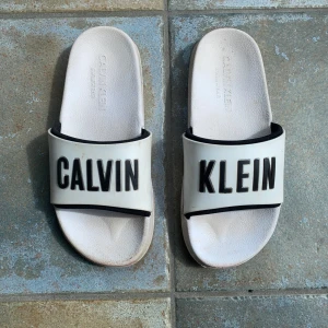 Vita sandaler från Calvin Klein - Snygga vita sandaler från Calvin Klein med svart logga över foten. De har en klassisk slip-on design och är tillverkade i syntetmaterial, perfekta för sommardagar eller chill hemma. Riktigt clean look med svart-vit kontrast. Ny pris är runt 1100