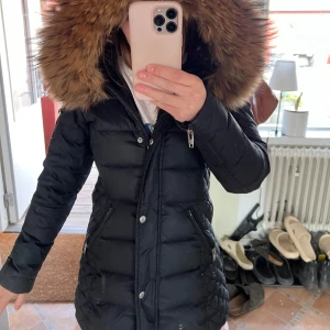 Svart dunjacka Rockandblue med päls - Säljer en svart lång dunjacka från Rockandblue med stor fluffig fuskpäls på huvan. Jackan har quiltade detaljer, dragkedjor på fickorna och en badge på ärmen. Perfekt för kalla dagar när du vill vara både varm och snygg.