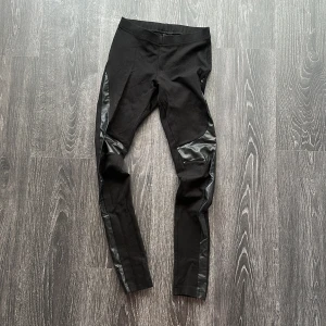 Svarta leggings med skinnimitation - Svarta leggings med snygga detaljer i skinnimitation längs sidorna och knäna. Tight passform och hög midja, perfekta för en edgy och trendig look. Mjukt material som sitter skönt hela dagen.
