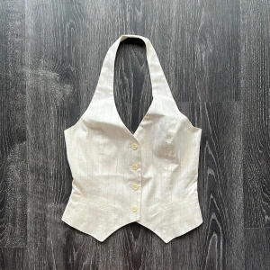 Vit randig omlottväst med knappar - Snygg vit väst med tunna ränder och omlottband i ryggen. Djup v-ringning och breda axelband som ger en cool halterneck-look. Västen har fyra knappar framtill och är sydd i ett lätt, lite glansigt tyg som känns lyxigt.