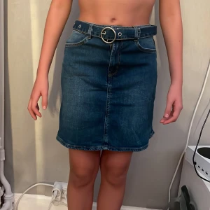 Mörkblå jeanskjol med bälte - Snygg mörkblå jeanskjol med coolt bälte och silverfärgat ring-spänne. Kjolen har klassiska fickor både fram och bak samt markerade sömmar. Perfekt för dig som gillar en enkel men trendig look.