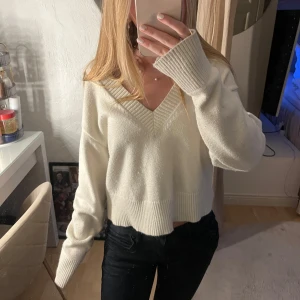 Vit croppad stickad tröja V-ringad - Superfin vit stickad tröja med croppad passform och djup v-ringning. Tröjan har breda ribbade muddar vid ärmslut och nederkant, vilket ger en snygg och avslappnad look. Perfekt att styla med jeans eller kjol för en trendig vibe.