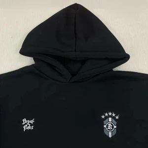 Svart hoodie från Brag Rides - Svart hoodie från Brag Rides med broderad logga och emblem på bröstet. Klassisk känguruficka framtill, huva och långa ärmar. Perfekt för dig som gillar streetwear och enkel stil. Materialet känns mjukt och bekvämt, passar till chill dagar.