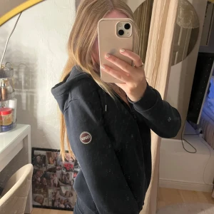 Svart hoodiejacka från Colmar - Svart hoodiejacka från Colmar med dragkedja framtill och huva med snörning. Jackan har ribbade muddar och två fickor på sidorna. Klassisk och enkel design med Colmar-logga på ärmen. Perfekt för chill dagar och streetstyle.