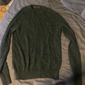 Mörkgrön merinoullströja Morris XS - Snygg mörkgrön tröja från Morris i 100% merinoull. Klassisk rund halsringning och diskret broderad logga på bröstet. Långärmad modell med ribbade muddar vid ärmslut och nederkant. Perfekt för dig som gillar stilrena och bekväma plagg.