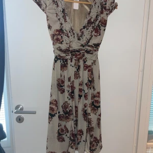 Blommig långklänning från Mango Suit - Supersöt långklänning från Mango Suit i beige med stora rosbruna blommor. Djup v-ringning med volangdetaljer och markerad midja. Korta ärmar och mjukt, följsamt material i 100% polyester. Perfekt för dig som gillar romantisk och bohemisk stil.