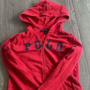 Ralph Lauren hoodie - Röd hoodie från Polo Ralph Lauren med stor huva och dragkedja framtill. Tröjan har mörkblå POLO-tryck över bröstet och är tillverkad i mjuk bomull. Perfekt för en chill och sportig look.