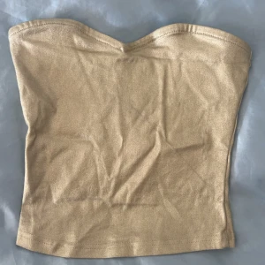 Guldig bandeau tubtopp - Säljer en snygg guldig bandeau tubtopp med glansig finish. Toppen är axelbandslös och har en enkel, stilren design som sitter tight mot kroppen. Perfekt att styla till jeans eller kjol för en trendig look.