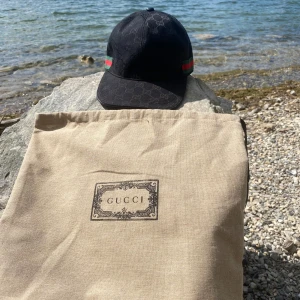 Svart Gucci keps med GG-mönster - Exklusiv svart keps från Gucci med diskret GG-monogram över hela kepsen. Klassisk böjd skärm och justerbar rem bak. Sidorna har den ikoniska gröna och röda randen. Tillverkad i bomull och levereras med originalpåse och tags.