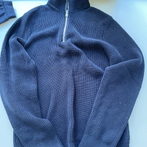 Mörkblå stickad half zip från East West - Mörkblå stickad tröja från East West i 100% bomull. Tröjan har hög krage och half zip-dragkedja framtill. Ribbstickad struktur och långa ärmar ger en clean och stilren look. Perfekt för lager-på-lager under kyliga dagar.