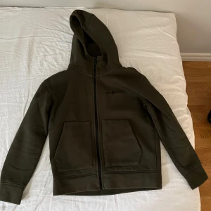 Mörkgrön hoodie från Sail Racing - Säljer en mörkgrön hoodie från Sail Racing med dragkedja och två stora fickor framtill. Tröjan har huva och är tillverkad i en skön blandning av bomull och polyester. Perfekt för kyliga dagar och har en clean look utan mönster.