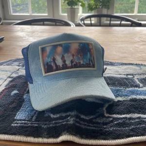 Blå truckerkeps från Maggiore - Snygg blå truckerkeps från Maggiore med mesh på sidorna och bak, samt ett coolt tryck framtill med konsertmotiv. Kepsen har böjd skärm och metallogo på sidan. Perfekt för dig som vill sticka ut med en unik accessoar.